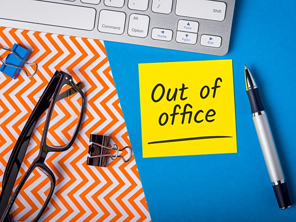 desk with yellow post-it note saying 'out of office' alongside keyboard, pen and reading glasses